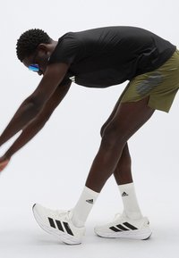 Μαύρο αθλητικό μπλουζάκι, πράσινο σορτς με γκρι λεπτομέρειες, λευκά αθλητικά παπούτσια με μαύρες ρίγες και μπλε γυαλιά ηλίου. Άνδρας σε θέση διάτασης.