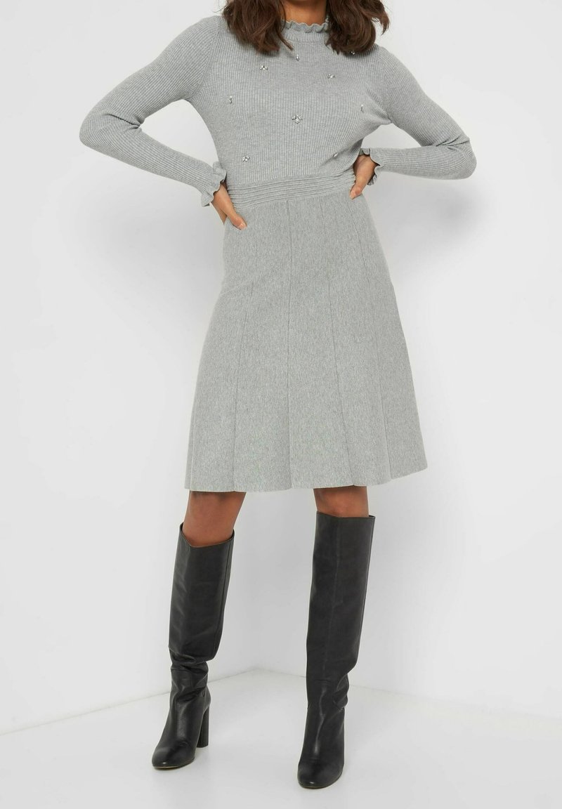 Haut côtelé gris à manches longues avec des accents en perles, associé à une jupe plissée grise et des bottes noires à talons montant jusqu'aux genoux. Texture lisse, design ajusté.