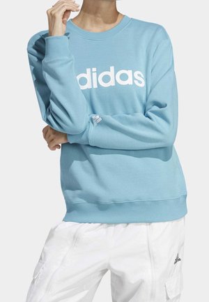 Personne portant un sweat Adidas bleu clair avec un logo blanc et un pantalon cargo Adidas blanc, bras croisés et main près du visage.