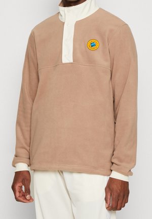 Sudadera de forro polar marrón con un alto cuello blanco y un parche redondo y colorido en el pecho. Mangas largas con puños ajustados.