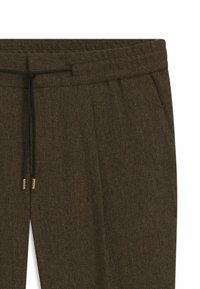 Bruine visgraatbroek met plooien, voorzien van een platte tailleband en een zwarte trekkoord. Messing aglets accentueren de uiteinden van het trekkoord.