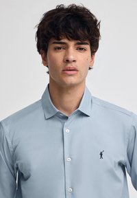 Giovane uomo con capelli ricci scuri che indossa una camicia azzurro chiaro con bottoni, caratterizzata da un piccolo logo ricamato sul petto.