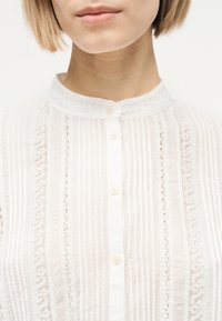 Γυναίκα που φοράει λευκό μπλουζάκι με δαντέλα στα τελειώματα, κάθετες πιέτες και κουμπιά, με ορατό λαιμό και κάτω μέρος προσώπου με ουδέτερη έκφραση.