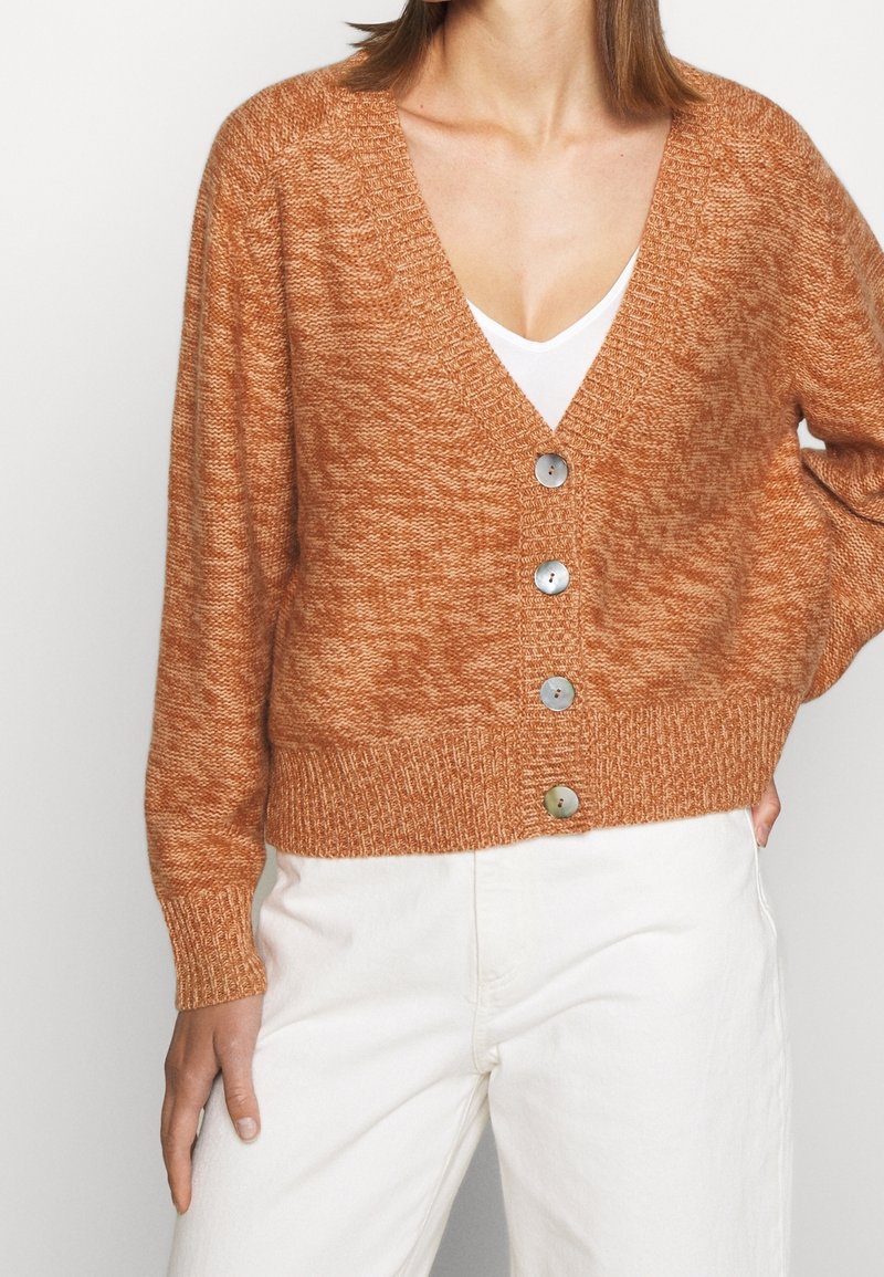 Žena ve zrezivě hnědém pleteném svetru s čtyřmi velkými knoflíky přes bílý top a bílé kalhoty, stojící před bílým pozadím.