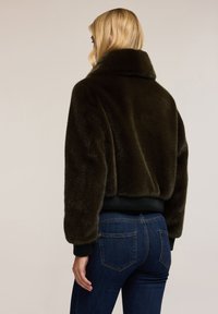 Blond kvinna som bär en mörkbrun faux fur kort jacka med ribbade manschetter och nederkant, i kombination med mörkblå denimjeans, vänd bort.