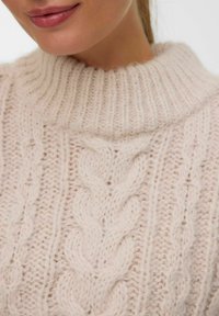 Gros plan sur une personne portant un pull en tricot torsadé rose clair avec un col haut à côtes et une texture douce.