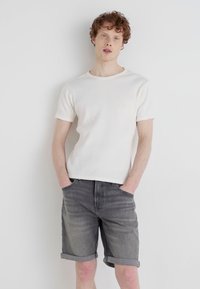 Bela rebrasta kratka majica s kratkimi rokavi v kombinaciji s sivo denim kratkimi hlačami, ki imajo zvit rob in žepke, stojijo pred enostavno ozadje.