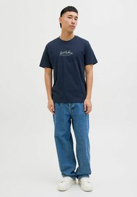 Tricou din bumbac de culoare bleumarin, cu mâneci scurte și gât rotund, având text alb pe piept; asortat cu blugi din denim de culoare albastră deschisă și adidași albi.
