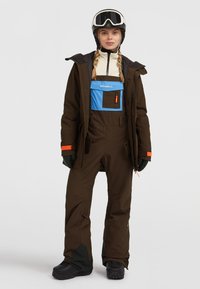 Bruine geïsoleerde skijack met een blauwe zak, zwarte handschoenen en skibril. Heeft oranje accenten en stevige zwarte sneeuwlaarzen.