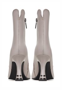 Bokáig érő bézs bőr csizma lekerekített orral, kontrasztos fekete talppal és fényes fémes kiemelésű tűsarokkal. Cipzáras záródással rendelkezik.