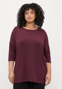 Camisola larga bordeaux com decote redondo e mangas três quartos, feita de um tecido suave. Bainha ligeiramente curva para um ajuste descontraído.