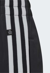 Czarne spodnie sportowe z szaro-białymi paskami, teksturowanym pasem w talii oraz małą etykietą "Koniec z odpadami plastikowymi" z boku.