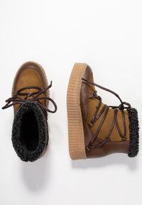Botas de tobillo marrones y tostadas con una suela de goma texturizada, forro de lana y cordones marrones. Se pueden ver materiales de ante y cuero.