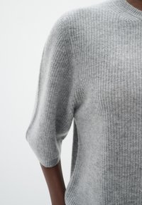 Šedý ribbed pletený svetr s uvolněným střihem a krátkými rukávy. Hladký povrch s jemnými variacemi odstínů po celém povrchu.