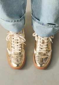 Goldfarbene Metallic-Sneaker mit weißen Akzenten, strukturierter Oberseite und beigen Schnürsenkeln, kombiniert mit hochgekrempelten hellblauen Jeans.