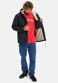 Černá nepromokavá bunda s béžovým fleece podšívkou, otevřená vpředu, s kapucí na stažení, nošená přes červené tričko a modré džíny. Černé boty.