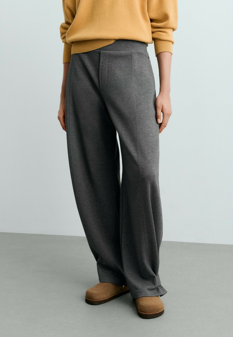 Personne portant un pantalon gris ample, un pull beige et des chaussures marron à enfiler, debout sur un sol gris devant un mur clair uni.