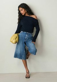 Navy off-shoulder långärmad topp i kombination med ljusblå vida jeansculottbyxor och svarta sandaletter med remmar, bärande på en gul handväska.