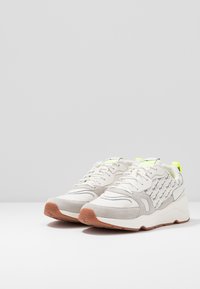 Sneakers na sznurówki z białej tkaniny i szarego zamszu, z jaskrawym żółtym uchwytem z tyłu oraz subtelnym nadrukiem logo na bokach.