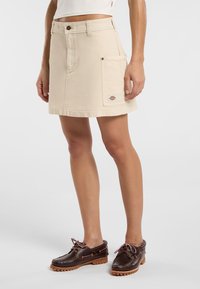 Beige denimkjol med rak passform, med sidoficka och en logopatch. Bärs med bruna snörskor och en vit topp.