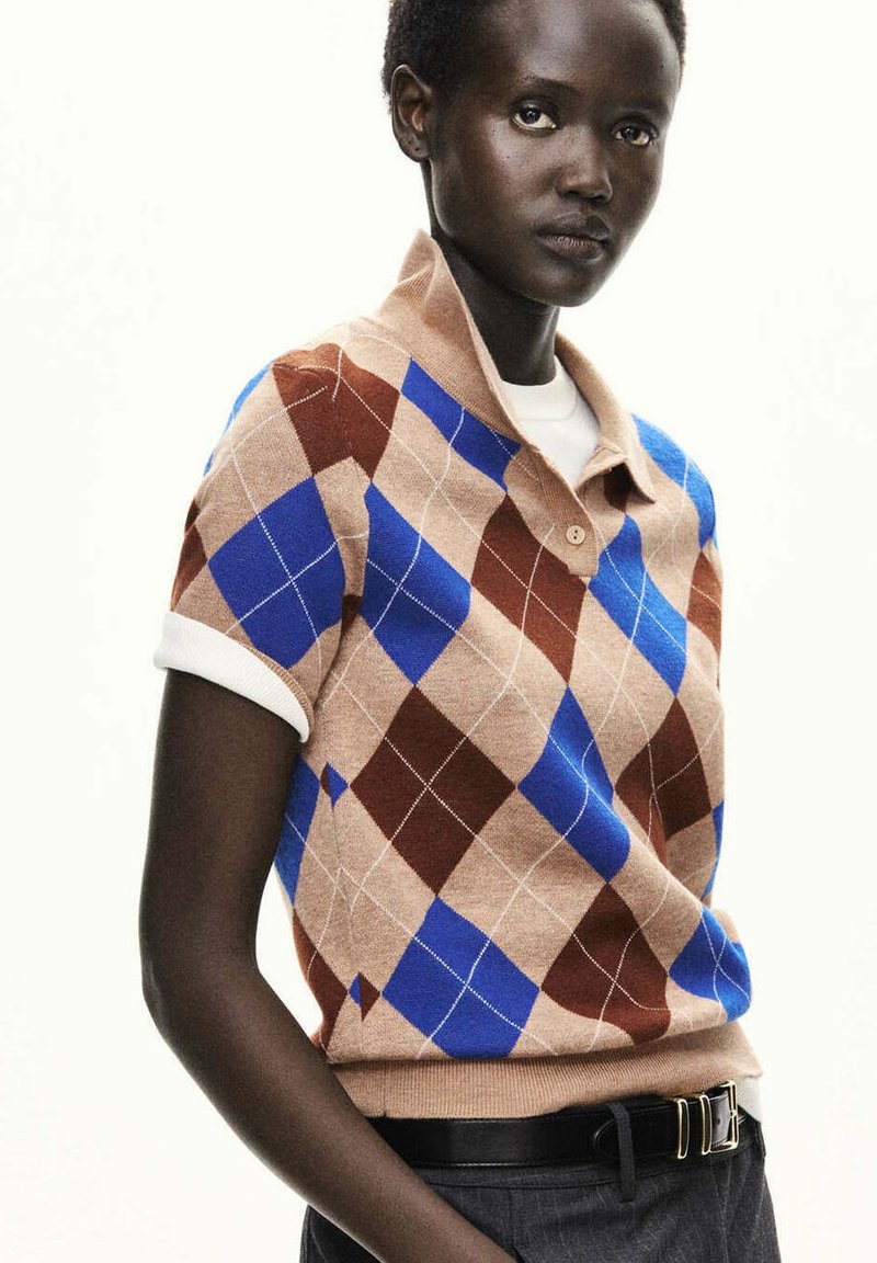 Personne portant un pull à manches courtes à motif losange beige, bleu et marron avec un col blanc, une ceinture noire et un pantalon foncé, sur un fond uni.