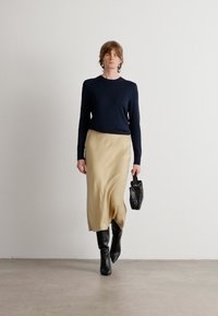 Marinegenser, beige satengskjørt og svarte knelengde støvler. Modellen holder en liten svart teksturert veske. Enkel bakgrunn.