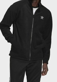 Svart fleecejacka med hel dragkedja, långärmad, ribbade muddar och sidofickor. Har en vit Adidas-logotyp på vänster bröst.