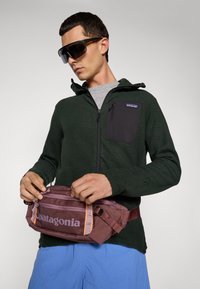 Vīrietis ar melnbaltām saulesbrillēm, tumši zaļā zip-up jakā ar krūšu kabatu, zilajos šortos un noregulē maroon Patagonia jostas somu.