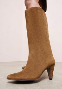 Cizme înalte din suede maro, cu vârf ascuțit și toc subțire, având o textură netedă și o siluetă elegantă.