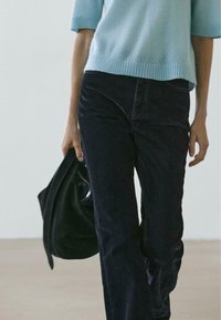 Maglione a maniche corte di colore azzurro chiaro abbinato a pantaloni svasati in denim scuro. Il look è completato da una giacca di pelle nera tenuta in mano.