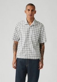 Camisa de manga corta en blanco con un patr�ón geométrico en gris. Presenta un cuello y un corte holgado, combinada con pantalones de mezclilla de un azul oscuro.