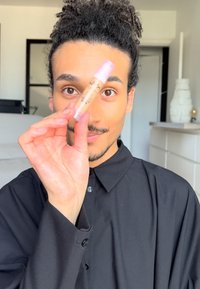Lucidalabbra trasparente in un tubo trasparente con un tappo rosa. La persona lo tiene sollevato contro uno sfondo chiaro, indossando una camicia nera con bottoni.
