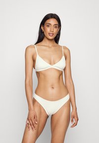 Conjunto de bikini creme com um triângulo com textura e bordas rendadas, acompanhado de cueca de bikini de cintura baixa com acabamentos decorativos.