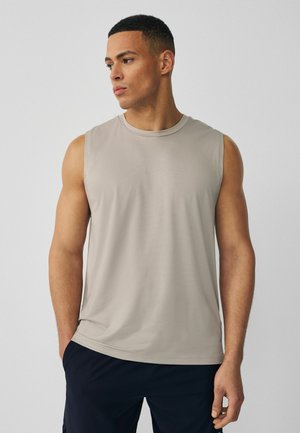 Mann trägt ein ärmelloses hellgraues Sportshirt und dunkle Shorts, schaut seitlich und hat die Hände in den Taschen, vor einem einfarbigen Hintergrund.