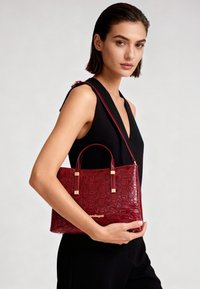 Rode leren handtas met reliëf en een gestructureerde vorm, voorzien van gouden metalen accenten en twee handvaten, vastgehouden tegen een zwart kledingstuk.