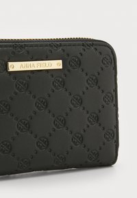 Portefeuille zippé noir avec des motifs circulaires en relief, plaque de marque dorée portant l'inscription "ANNA FIELD" et surface texturée pour un détail tactile.