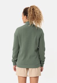 Felpa verde, con texture, a collo alto e con polsini a coste. Indossata con pantaloni corti beige, che mostrano un look casual e rilassato.