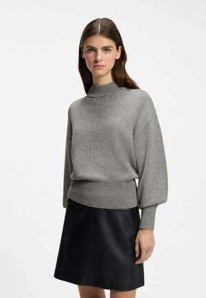 Junge Frau mit langen braunen Haaren, die einen grauen Strickpullover und einen schwarzen Lederrock trägt, steht vor einem weißen Hintergrund.