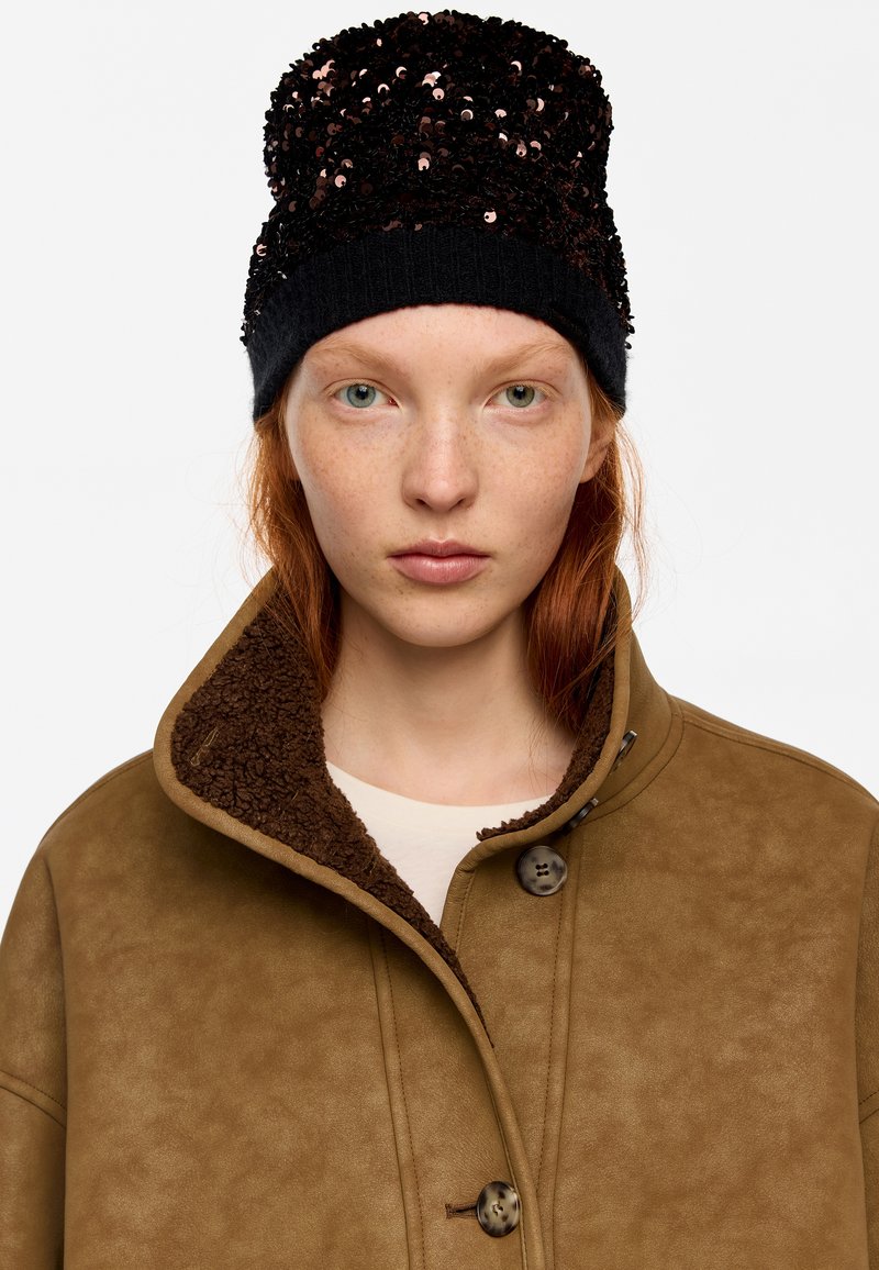 Gorro negro con lentejuelas y parte superior texturizada, combinado con una chaqueta marrón de ante sintético que presenta un cuello de felpa y dos botones.