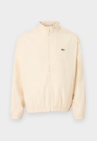 Μπεζ μπουφάν με φερμουάρ, ελαστικά μανσέτες και τελείωμα, ψηλό γιακά και μικρό πράσινο λογότυπο κροκόδειλου στο αριστερό στήθος.