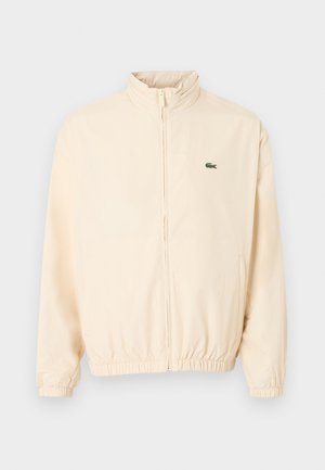 Bež jakna z zadrgo, elastičnimi manšetami in spodnjim robom, visokim ovratnikom ter majhnim zelenim logotipom krokodila na levi strani prsnega koša.