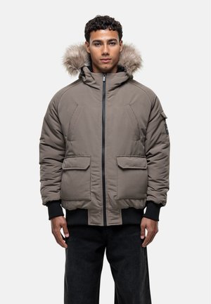 Brown puffer jacket with a fur-lined hood, two front pockets, and ribbed cuffs. Features a central zip closure and a patch on the sleeve.