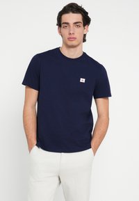 Tmavomodré bavlnené tričko s okrúhlym výstrihom a krátkymi rukávmi. Na ľavej strane na hrudi sa nachádza malá obdĺžniková biela etiketa s červeným logom.