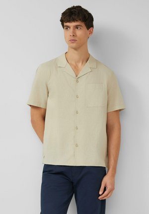 Homme portant une chemise beige à manches courtes boutonnée avec une poche poitrine et un pantalon bleu foncé, debout devant un mur uni gris clair.