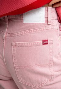 Růžové džínové kalhoty mají klasický design s pěti kapsami, bílou koženou zadní štítku a červenou značkou na pravé zadní kapse.