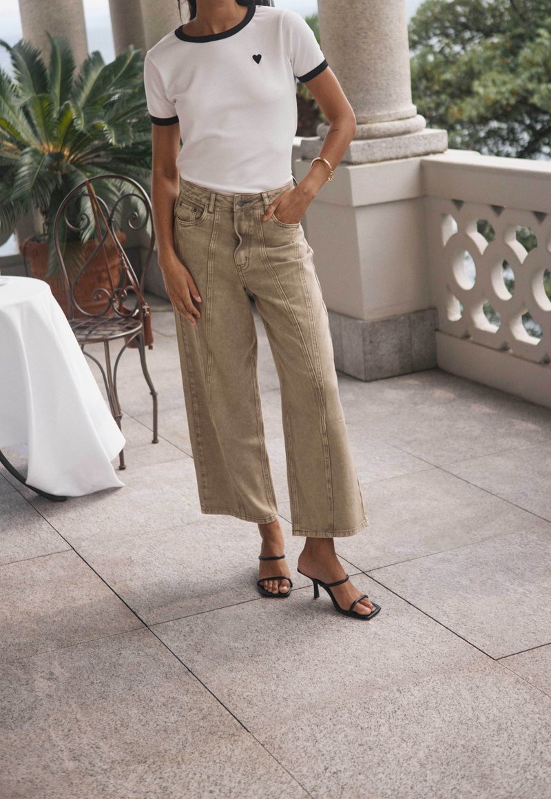 Femme portant un pantalon beige à jambes larges, un t-shirt blanc avec un liseré noir et un détail en forme de cœur, et des talons noirs à brides, debout sur une terrasse en pierre.