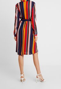 Robe rayée multicolore avec des bandes rouges, bleu marine, orange et blanches, à manches longues et taille nouée. Associée à des sandales à talons blancs.