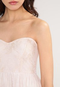 Sukienka bez ramiączek w jasnym beżu z koronkowymi detalami kwiatowymi na górze oraz przezroczystym materiałem na spódnicy, ukazująca miękką teksturę i subtelny połysk.