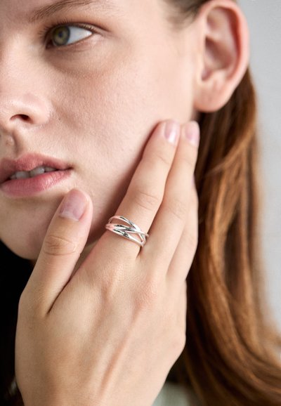 Bague en argent avec un design entrelacé, comportant des sections lisses et texturées. Portée sur une main touchant le visage, mettant en valeur un savoir-faire détaillé.