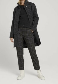 Lungo cappotto di lana grigio scuro sopra un maglione nero, abbinato a pantaloni a quadri neri e grigi e sneakers alte bianche.